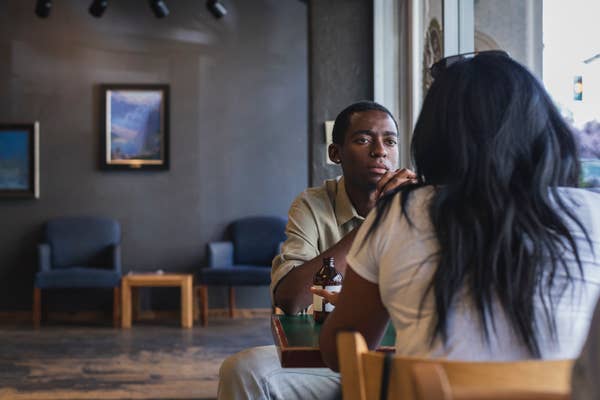 A person and a woman sit across from each other at a cafe table, engaged in conversation, with artwork and seating visible in the background