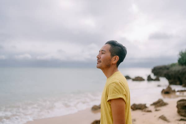 A person stands on a rocky beach, gazing thoughtfully at the sea, wearing a short-sleeved shirt