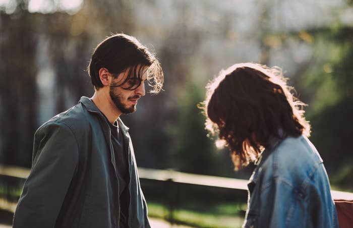 Two people outdoors, both looking down, wearing casual jackets. Background includes trees and natural light. Faces are not clearly visible