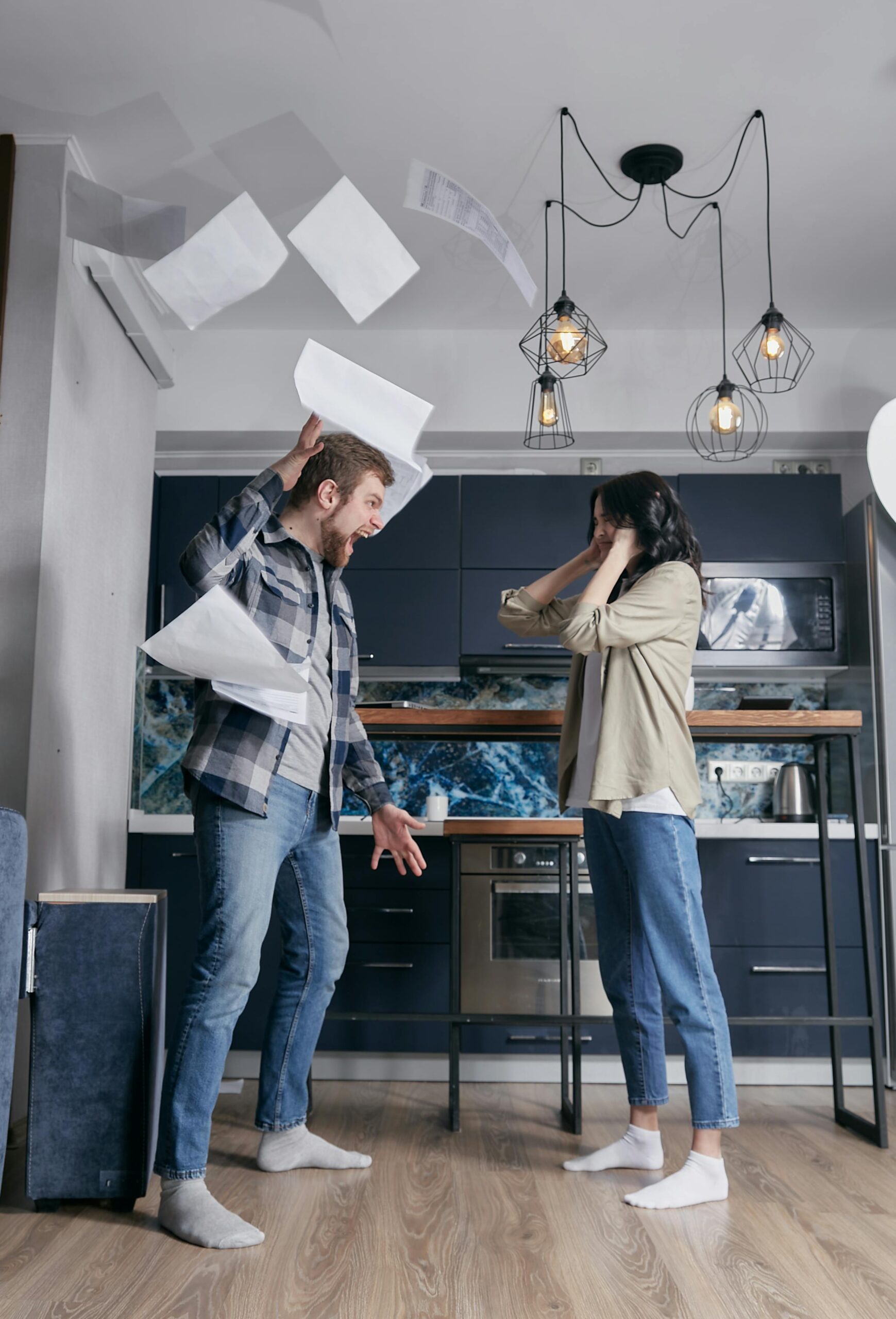 A man and woman argue in a kitchen with papers flying around.