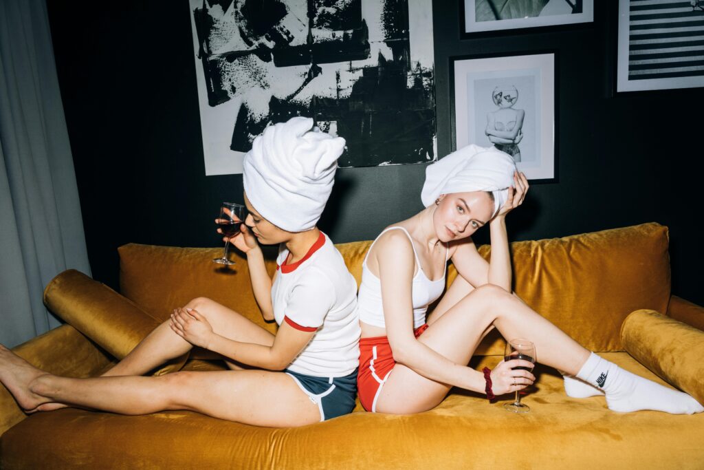 Two young women enjoy a pajama party, sipping wine on a stylish yellow sofa.