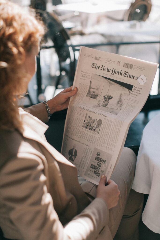 A woman with blond hair reads The New York Times newspaper indoors during the day.